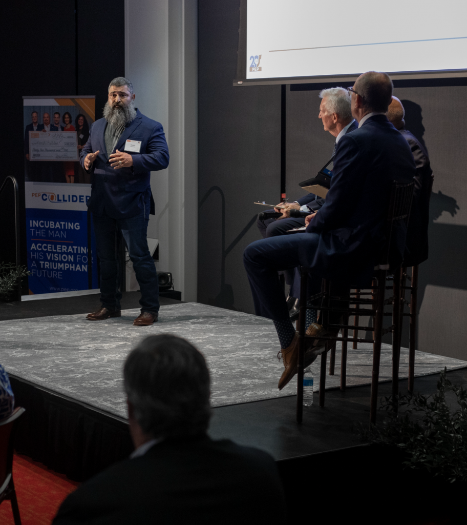 PEP graduate in a suit presents on stage during a business pitch event while a panel of judges listens attentively.